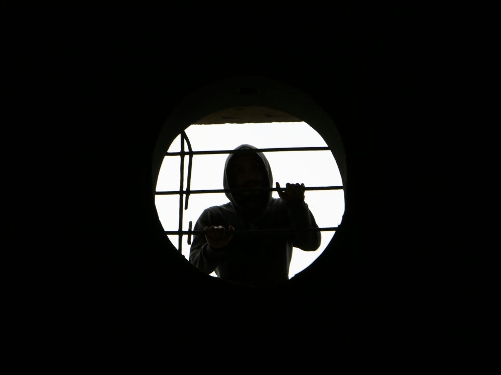 Silhouette of a person in a hoodie behind a round, barred window creating a mysterious scene.